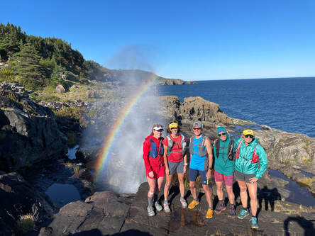 5 people on a trail running trip and in bright running gear posing together by the edge of a cliff with a rainbow in the background